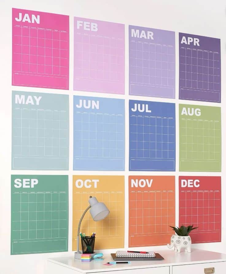 Douze feuilles de calendrier mensuel colorées, accrochées au mur au-dessus d'un bureau, servant de calendrier éditorial pour les réseaux sociaux.