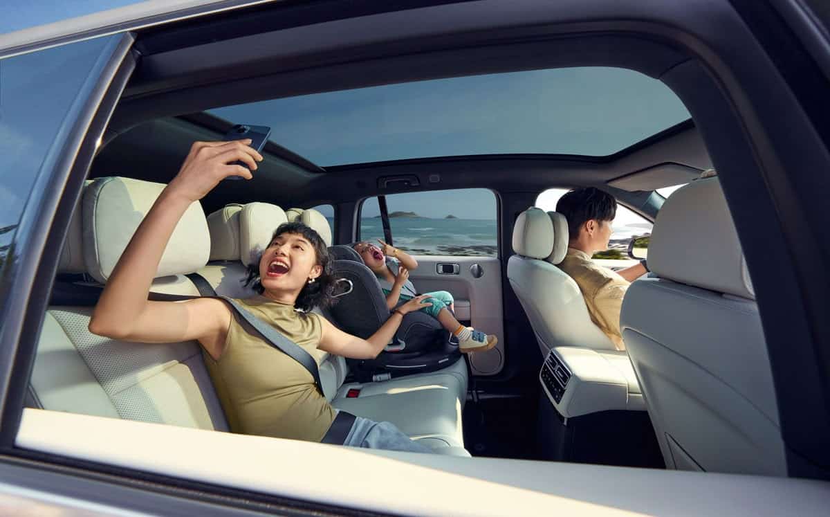 Une femme prend un selfie dans le cadre d'une campagne de marketing d'influence sur la banquette arrière d'une voiture, tandis qu'un enfant est assis dans un siège auto et qu'un homme conduit.